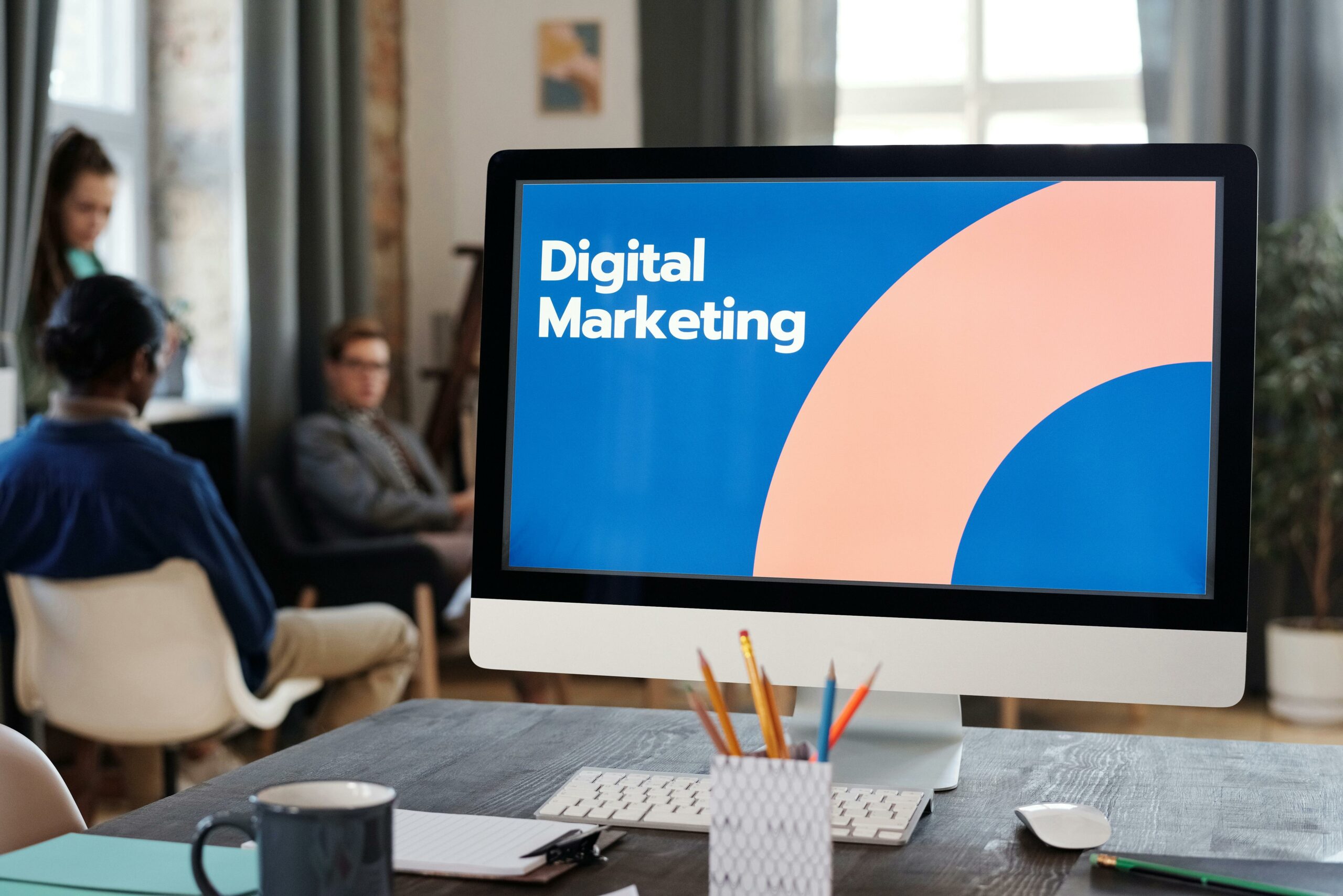 Digital Marketing on Screen Computer screen on a desk displaying 'Digital Marketing,' with a blurred background featuring three individuals – two seated on chairs and one standing. Shallow depth of field emphasizes the focal point on the screen and workspace.